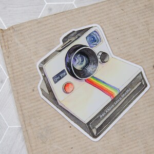 Pu&ograve; includere: Un'illustrazione ad acquerello di una vintage Polaroid Land Camera con una striscia arcobaleno sul davanti. La fotocamera &egrave; bianca con accenti neri e dice "One Step" sul davanti.