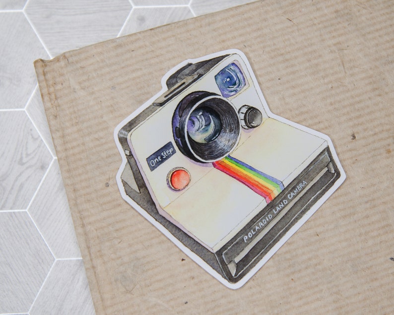 Pu&ograve; includere: Un'illustrazione ad acquerello di una vintage Polaroid Land Camera con una striscia arcobaleno sul davanti. La fotocamera &egrave; bianca con accenti neri e dice "One Step" sul davanti.