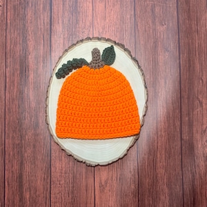 May include: Orange crocheted pumpkin hat with green leaves on a wooden background.