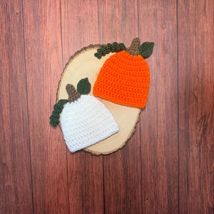 May include: Two crocheted hats shaped like pumpkins, one in bright orange and one in white. Each hat features a brown stem, a dark green leaf, and a small dark green curly vine attached to the top, resting on a light-colored wooden slice.