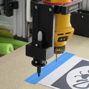 May include: A CNC machine with a yellow and black router bit is positioned over a paper stencil. The machine is mounted on a metal frame. Blue tape secures the stencil. The stencil features a black and white design.