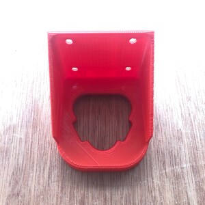 May include: A red 3D printed plastic bracket with four holes for mounting. The bracket has a cutout in the centre.
