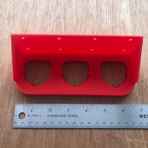 May include: A red plastic 3D printed wall mount with three circular cutouts. The mount is 15 cm long and has a ruler underneath it.