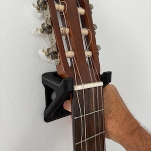 May include: Acoustic guitar headstock and neck, mounted on a black wall hanger. The guitar has a dark brown wood finish, white tuning pegs, and silver strings. The fretboard is a darker shade of brown.
