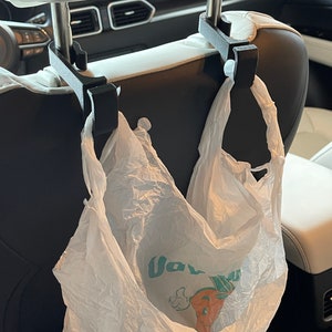 May include: Two black plastic hooks are hanging from the headrests of a car seat. A white plastic shopping bag with a green and orange logo is hanging from the hooks.