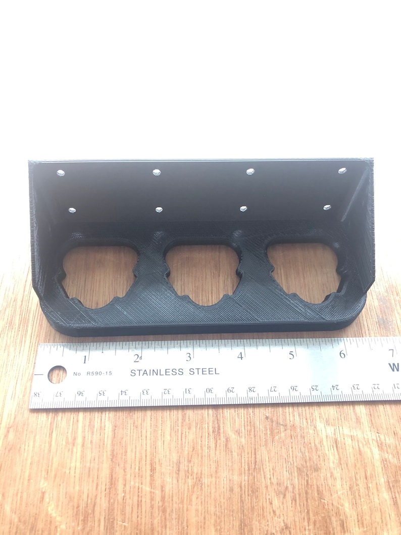 May include: A black plastic 3D printed shelf with three circular cutouts. The shelf is designed to hold round objects, such as cans or bottles. The shelf is measured with a ruler, showing it is approximately 15 cm long.