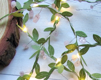 May include: A string of LED fairy lights featuring a flexible silver wire intertwined with artificial green leaves and small, pale pink tumbled stones. The lights emit a warm yellow glow, creating a vine-like appearance throughout the strand.