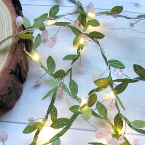 May include: A decorative garland featuring green fabric leaves and small pink gemstones. Tiny warm white LED lights are interspersed throughout the garland. The garland is arranged on a light-colored wooden surface.