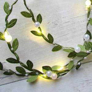 May include: A string of fairy lights with green leaves and white pearl-like beads. The lights are strung on a wire and are lit up.