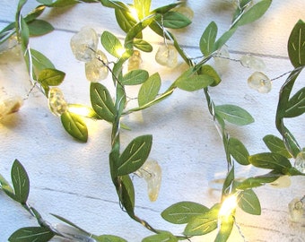 May include: A decorative string of lights featuring thin silver wire intertwined with green fabric leaves and small, translucent yellow crystal-like stones. The lights emit a warm glow, illuminating the foliage and stones along the entire length.