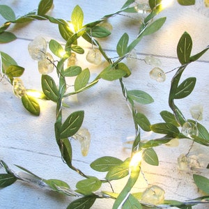 May include: A string of fairy lights with green leaves and clear glass beads. The lights are strung on a wire and are lit up. The lights are on a white wooden surface.