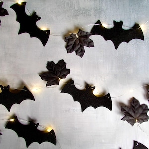 May include: A string of decorative lights featuring black paper bat silhouettes and dark fabric maple leaves. The thin wire connects each element, with small warm white LED lights positioned behind the bats and leaves to create a glowing effect.