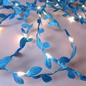 May include: A string of decorative lights featuring bright blue fabric leaves attached to a thin, flexible silver-coloured wire. Small, warm white LED bulbs are interspersed throughout the vine-like structure, illuminating the blue foliage.