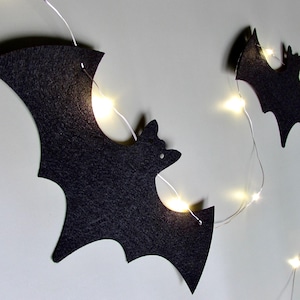 May include: A string of black felt bat-shaped cutouts connected by a thin silver wire featuring small, glowing warm white LED lights. The bats are arranged in a sequence, with each cutout displaying a distinct silhouette of a flying bat.