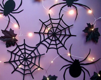 May include: A string of LED fairy lights featuring black die-cut decorations including spiders, spider webs, and maple leaves. The thin silver wire connects the warm white glowing lights, which are interspersed among the black shapes.
