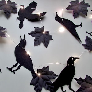May include: A string of decorative lights featuring black felt cutouts of birds and maple leaves. The thin silver wire connects the small, glowing white LED bulbs, which are interspersed between the dark bird silhouettes and leaf shapes.
