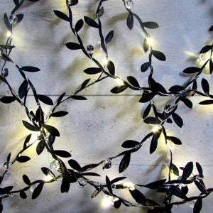 May include: A decorative string of lights featuring thin, flexible wire intertwined with small, matte black artificial leaves and clear, spherical beads. The integrated LED lights emit a warm, soft glow throughout the vine-like structure.