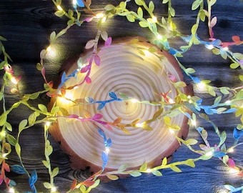 May include: A string of decorative fairy lights featuring artificial vine leaves in shades of green, blue, pink, and yellow. The flexible wire is intertwined with small, warm white LED bulbs that emit a soft glow throughout the foliage.