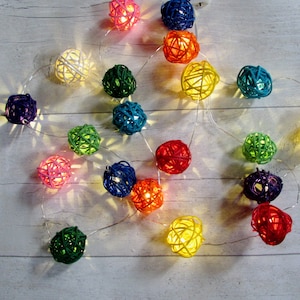 May include: A string of decorative lights featuring spherical shades made of woven rattan-like material in various colors including white, pink, yellow, orange, red, green, blue, and purple, connected by a thin, flexible silver-toned wire.