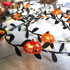 May include: A string of decorative lights featuring small orange plastic jack-o'-lanterns interspersed with black fabric leaves. The pumpkin-shaped covers are illuminated from within by warm white LED lights, creating a glowing effect along the vine.