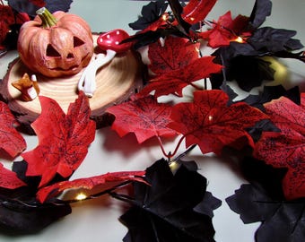May include: A string of artificial maple leaves in red and black colors, interspersed with small warm white LED lights. A carved orange pumpkin, a small red and white mushroom figurine, and a tiny fox figure sit on a round wooden slice.