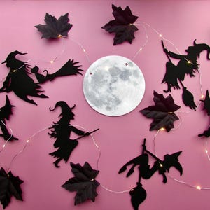 Könnte beinhalten: Eine dekorative Lichterkette mit schwarzen Silhouetten von Hexen auf Besen, dunkelvioletten Ahornblättern und einem zentralen runden Mond-Ausschnitt. Der dünne silberne Draht ist mit kleinen, warmweißen LED-Lichtern durchzogen.