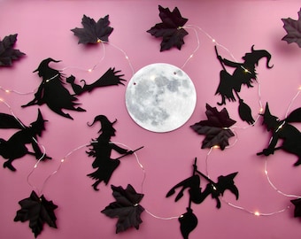 May include: A decorative string light set featuring black silhouettes of witches on broomsticks, dark purple maple leaves, and a central circular moon cutout. The thin silver wire is illuminated by small, warm white LED lights throughout.
