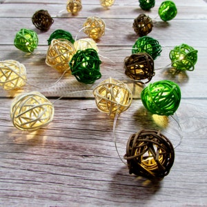 May include: A string of lights with white, brown, and green woven ball decorations. The lights are strung on a white wire and are hanging on a wooden surface.