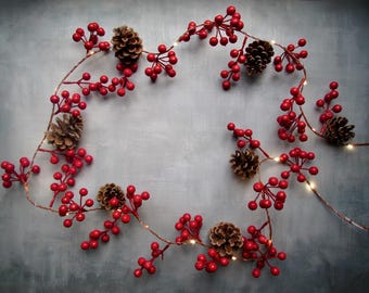 May include: A decorative garland featuring clusters of bright red berries and brown pinecones arranged along a thin, flexible wire string. Small, warm-toned LED lights are interspersed throughout the length of the garland, creating a glowing effect.