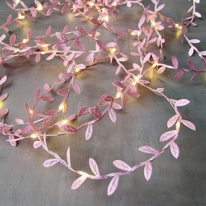 May include: A string of pink paper leaves with small, warm white LED lights. The leaves are arranged along a thin wire, creating a decorative garland. The lights are evenly spaced, providing a soft glow.