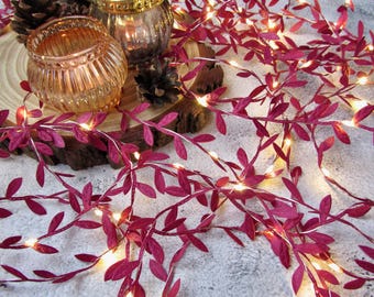 May include: A string of LED fairy lights featuring dark pink artificial leaves intertwined with the wire. Two ribbed glass candle holders sit on a circular wooden slice, accompanied by small brown pinecones nestled near the base of the lights.