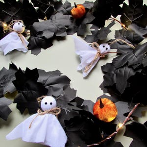 May include: A decorative garland featuring black artificial maple leaves, small white fabric ghost figures with black eyes, and miniature orange pumpkins. The arrangement is accented with thin twine bows and integrated warm white LED string lights.
