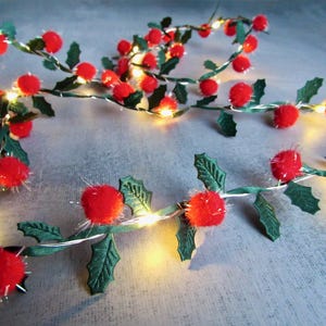May include: A Christmas garland with red pom-pom berries, green fabric leaves, and small, warm white lights. The garland resembles holly and is suitable for festive decorations. The lights are strung along a thin, flexible wire, perfect for draping.