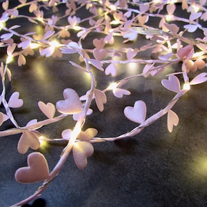 May include: A string of fairy lights featuring small, pale pink fabric hearts attached to a thin, flexible silver wire. The lights emit a warm, soft glow, illuminating the heart-shaped accents along the length of the decorative cord.