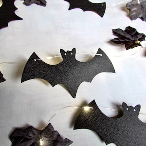 May include: A string of decorative lights featuring black paper bat cutouts and dark fabric leaves. The thin silver wire connects the individual bats and leaves, with small warm white LED bulbs positioned behind the cutouts to provide illumination.