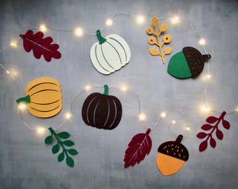 May include: A string of LED fairy lights decorated with felt shapes including pumpkins in white, yellow, and dark brown, green and red leaves, a yellow branch, and two acorns with brown caps and green or yellow bodies arranged in a festive display.
