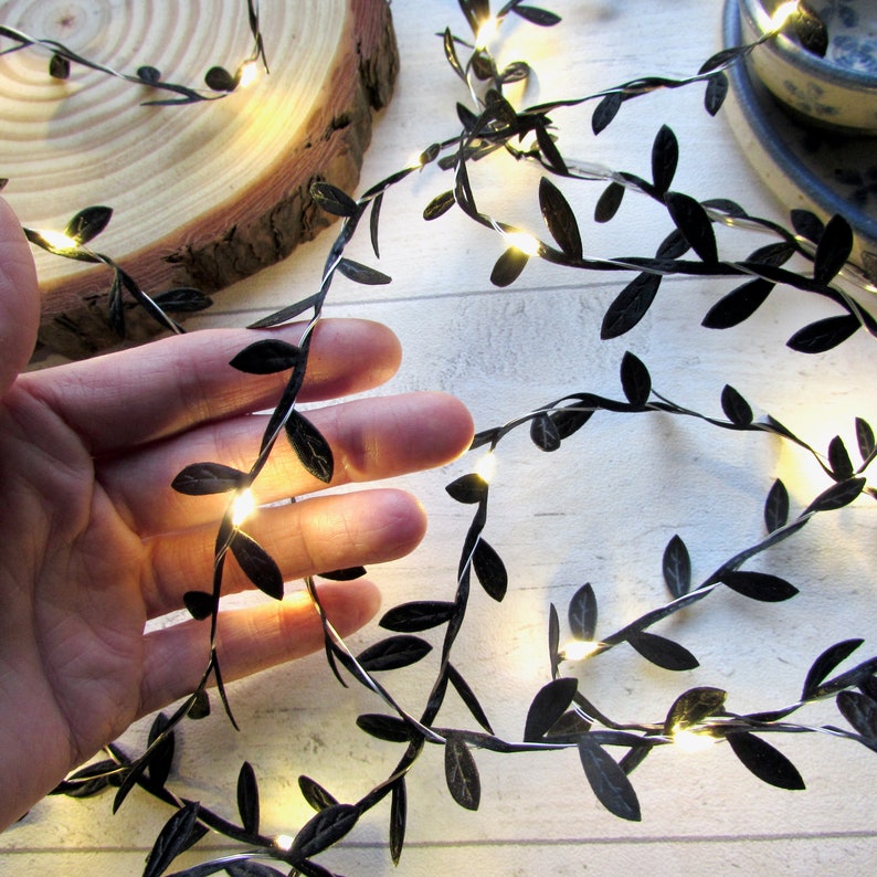 A hand holding the warm white LED fairy lights intertwined with black fabric mini leaf ribbon - demonstrating that the leaves are approx 1.5cm in length