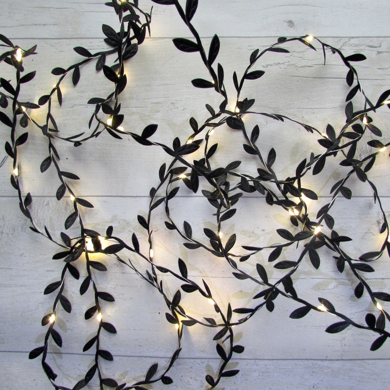 May include: A string of decorative fairy lights featuring thin, flexible wire intertwined with small, matte black artificial leaves. The strand is illuminated by numerous tiny, warm white LED bulbs positioned at intervals along the vine-like structure.