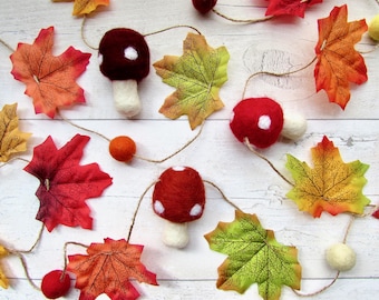 May include: A decorative garland featuring felted wool mushrooms in shades of red and brown with white spots, interspersed with artificial maple leaves in autumn colors of orange, red, and yellow, all strung together on a thin brown twine cord.