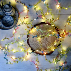 May include: A string of fairy lights with colorful leaf-shaped decorations. The lights are wrapped around a wooden slice and are next to a blue and white floral tea set. The leaves are in shades of green, pink, and blue.