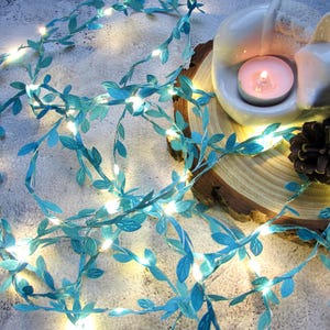 May include: A string of teal blue fairy lights with small leaf-shaped accents. The lights are wrapped around a wooden slice with a white ceramic figurine and a lit tea light candle.