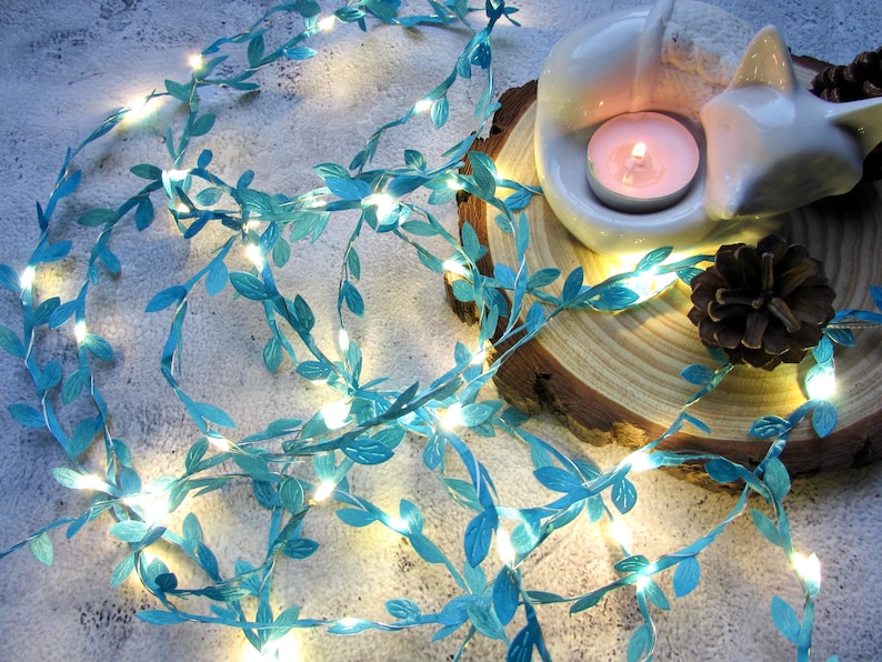 May include: A string of teal blue fairy lights with small leaf-shaped accents. The lights are wrapped around a wooden slice with a white ceramic figurine and a lit tea light candle.