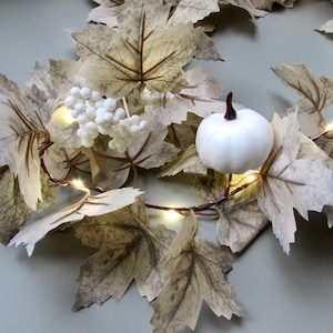 May include: A decorative garland featuring light beige maple leaves, clusters of small white berries, and a smooth white pumpkin with a dark brown stem. The arrangement is intertwined with thin copper wire string lights that emit a warm glow.