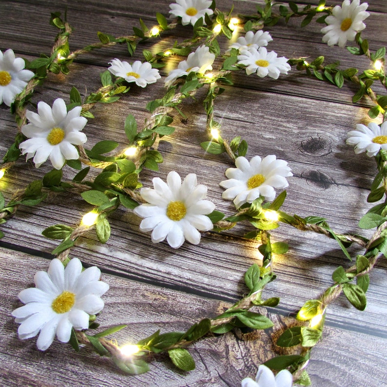 以下が含まれることがあります： 白いデイジーの花と緑の葉がワイヤーに巻き付けられたフェアリーライトのストリング。ライトは茶色の紐に結ばれています。
