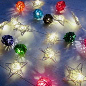 May include: A string of decorative lights featuring woven rattan spheres and star-shaped frames in various colors including red, blue, green, black, and white. Each element contains a small internal light that emits a warm glow.