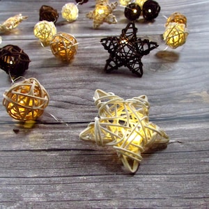 May include: A string of decorative lights featuring woven rattan spheres and star-shaped ornaments in natural beige and dark brown colours. Each piece contains a small internal light that emits a warm glow, connected by a thin, clear wire.
