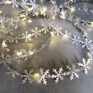 May include: A string of silver-colored snowflake-shaped ornaments connected by a thin wire, featuring integrated small warm white LED lights that illuminate the metallic cutouts, creating a continuous decorative garland of repeating snowflake patterns.