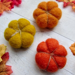 May include: Three needle-felted pumpkins in shades of mustard yellow, bright orange, and burnt orange. Each pumpkin features a rounded, segmented shape with visible twine wrapped around the exterior to define the sections of the wool fiber.