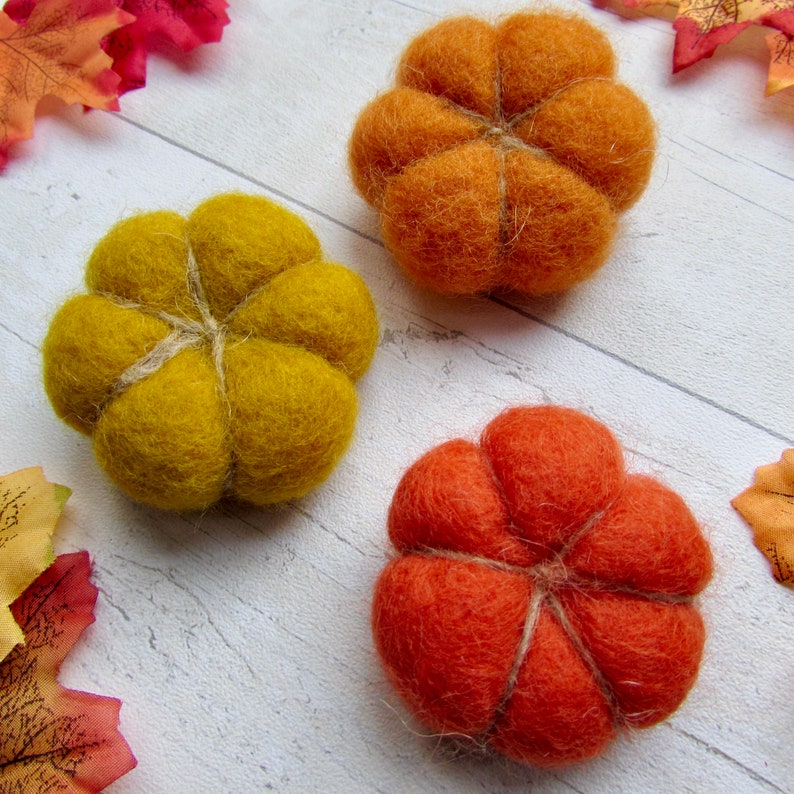 May include: Three needle-felted pumpkins in shades of mustard yellow, bright orange, and burnt orange. Each pumpkin features a rounded, segmented shape with visible twine wrapped around the exterior to define the sections of the wool fiber.