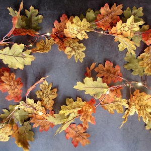 May include: A decorative garland featuring artificial oak leaves in shades of yellow, orange, green, and brown, interspersed with small, warm-toned LED fairy lights on a thin, flexible wire, creating a seasonal display of autumn foliage.